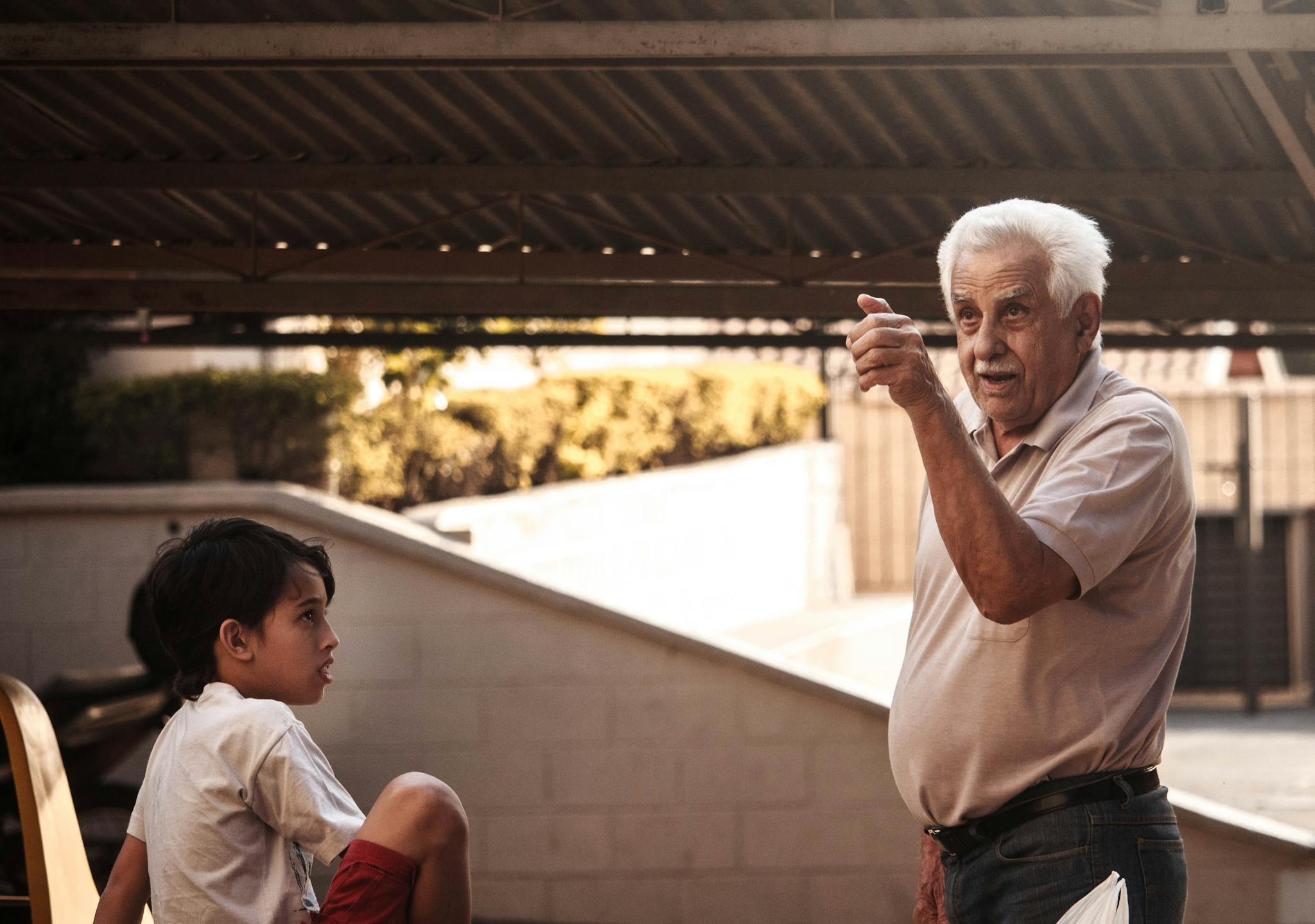 A senior adult gesturing and talking to a young boy outdoors under a canopy.